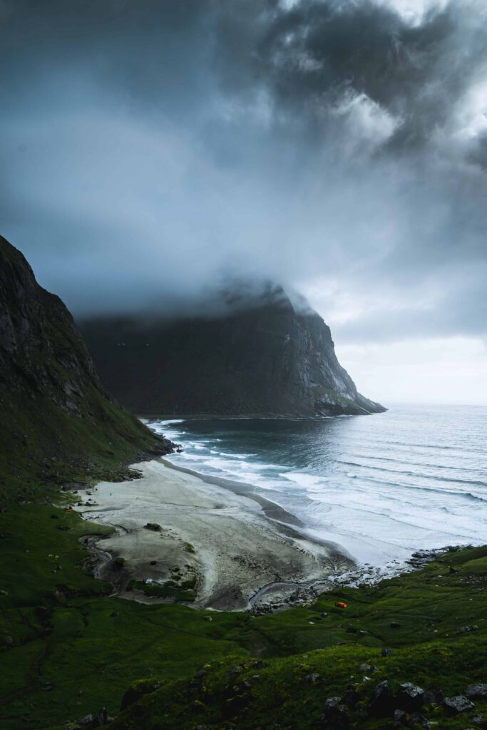 En ensam strand med tält vid foten av ett stort berg under en mörk, molnig himmel.